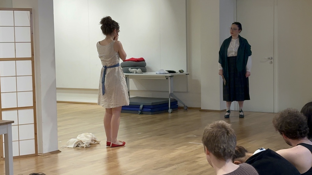 Two actors stand facing each other in mid-performance of 'Faith Hope Charity'. A few students sit in the audience watching the performance.